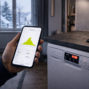 Intérieur de maison en hiver, smartphone affichant une courbe de production solaire, lave-vaisselle ou lave-linge programmé, lumière froide en journée